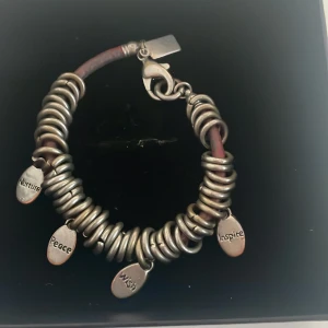 Silverfärgat armband med berlocker - Säljer ett coolt armband med silverfärgade ringar och berlocker med orden 'Nurture', 'Peace', 'Wish' och 'Inspire'. Armbandet har en robust design med en säker låsning. Perfekt för att ge en personlig touch till din stil!