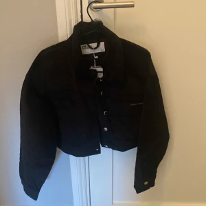 Svart Manchester jacka, midjekort - Säljer en snygg svart croppad jacka från Calvin Klein. Den har en cool design med knappar framtill och långa ärmar. Perfekt för en trendig look i höst eller vår. Passar bra till både vardag och fest!