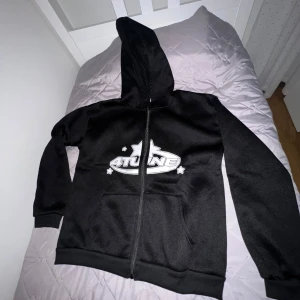 Svart hoodie med tryck - Säljer en svart hoodie med coolt tryck på framsidan. Den har en dragkedja och en stor ficka framtill. Perfekt för höst och vinter, och den är i bra skick. Passar både till vardags och chillkvällar. Hör av dig om du är intresserad!