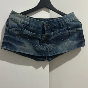 Blå jeansshorts med detaljer - Snygga blå jeansshorts i storlek XL med coola knapplås och glittriga detaljer framtill. Perfekta för sommardagar eller en avslappnad fest. De har en låg midja och klassiska bakfickor. Passar perfekt till en t-shirt eller linne för en avslappnad look.
