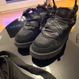 Svarta sneakers från UGG - Säljer ett par svarta sneakers från UGG i mycket bra skick. De har en snygg och robust design med tjocka snören och en bekväm sula. Perfekta för höst och vinter med sitt varma foder. Skorna är i syntetmaterial och har en cool, urban stil.