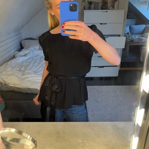 Svart blus med knytband  - Snygg svart blus med korta ärmar och dekorativa knappar vid axeln. Den har ett elegant knytband i midjan som ger en fin siluett. Perfekt för både vardag och fest! Vet dock inte vart den är ifrån då jag själv köte den begagnad och de inte står. Men bra skick iallafall!
