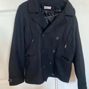 Svart kappa från H&M - Snygg svart kappa från H&M i bra skick. Den har en klassisk design med knappar framtill och dragkedjor på bröstfickorna. Perfekt för höst och vår, med en stilren look som passar både till vardags och fest. Storleken är pojke 170