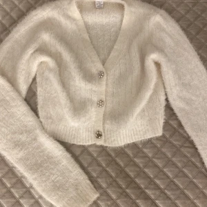 Vit fluffig kofta med pärlknappar - Superfin vit kofta i fluffigt material med v-ringning och glittriga knappar med pärlor. Det har åkt bort några pärlor från knapparna men man märker det knappt. Den är croppad och har långa ärmar, vilket gör den idealisk för både höst och vintern. ❣️