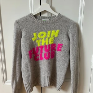 Grå kashmirtröja från From Future - Säljer en grå kashmirtröja från From Future med texten 'JOIN THE FUTURE CLUB' i neonfärger på framsidan. Tröjan har långa ärmar och ribbade muddar vid ärmslut och nederkant. Perfekt för en trendig look!