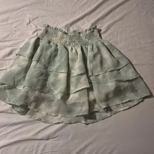 Grön volangkjol - Säljer en söt grön volangkjol med elastisk midja. Kjolen har ett mjukt, flödande tyg med flera lager som ger en fin volangeffekt. Kjolen är utav ett tunnt material, perfekt för sommaren.