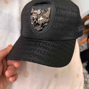Svart keps med metallisk detalj - Snygg svart keps från Philipp Plein med ett coolt mönster och en iögonfallande metallisk djurdetalj framtill. Perfekt för att ge din outfit en edgy touch.