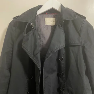 Svart trenchcoat från Banana Republic - Snygg svart trenchcoat från Banana Republic med knappar framtill och klassisk krage. Jackan har långa ärmar och ett stilrent snitt som passar perfekt för höst och vår. Perfekt för att ge en elegant touch till din outfit.