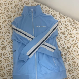 Ljusblå kofta från Palm Angels - Säljer en snygg ljusblå kofta från Palm Angels. Koftan har en dragkedja framtill och vita samt svarta ränder längs ärmarna. Perfekt för en sportig look med ribbade muddar och hög krage. Priset kan sänkas vid snabb affär . Har använt den ett par ggr. Pm vid snabb affär