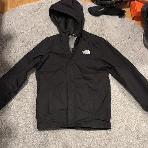 Svart vindjacka från The North Face - Säljer en svart vindjacka från The North Face med huva och dragkedja. Jackan har en stilren design med logotyp på bröstet. Perfekt för blåsiga dagar och lätt att packa med sig. Priset kan ändras vid snabb affär