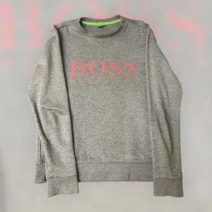 Grå sweatshirt från Hugo Boss - Snygg grå sweatshirt från Hugo Boss med röd logotyp på bröstet. Tröjan har en slim fit passform och är tillverkad i ett mjukt material. Perfekt för en stilren och bekväm look. Köpte den dyr och är äkta från själva Hugo boss 