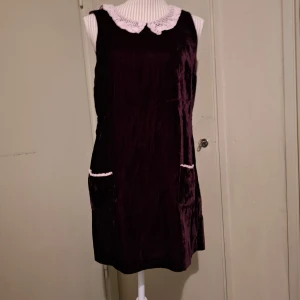 Mörklila sammet klänning med spetskrage - Säljer en elegant mörklila sammet klänning från My Style. Klänningen har en vacker vit spetskrage och är ärmlös. Den har en dragkedja i ryggen och två praktiska fickor framtill. Perfekt för en stilfull look!