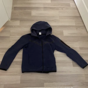 Mörkblå Nike tech Fleece - Säljer en mörkblå jacka från Nike med huva och dragkedja. Jackan har långa ärmar och en stilren design med svarta detaljer. Perfekt för en sportig look.