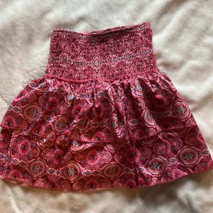 Mönstrad rosa kjol - Säljer en färgglad och mönstrad rosa kjol med volangdetaljer. Kjolen har en elastisk midja för bekväm passform och ett vackert paisleymönster i olika nyanser av rosa och rött. Perfekt för att liva upp garderoben och ha en varm sommardag. Insydda shorts. Aldrig använd så skicket är 10 av 10. Vid frågor skicka dm.