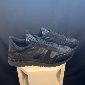 Svarta sneakers från Valentino Garavani - Snygga svarta sneakers från Valentino Garavani med en cool camouflage-design. Skorna har snörning och är tillverkade i en kombination av mocka och skinn. Perfekta för en stilren look. Äkta såklart! Som nya i skicket! Kan gå ned i pris vid snabb affär