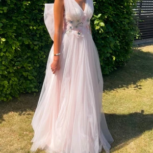 Ljusrosa långklänning med blommor - Elegant ljusrosa långklänning från Ida Sjöstedt med v-ringning och vackra blommiga detaljer i midjan. Klänningen har en stor rosett på ryggen och är ärmlös. Nypris: 11 000kr