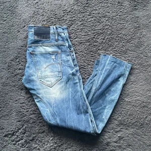 G-star jeans - Snygga blå jeans från G-Star med en sliten look och ljusa detaljer. De har en klassisk femficksdesign och en knappgylf. Perfekta för en avslappnad stil.