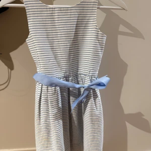 Randig klänning från Ralph Lauren - Barnklänning från Ralph Lauren i blått och vitt. Klänningen har en ljusblå knytning i midjan och knappar på baksidan. Perfekt för en somrig look!