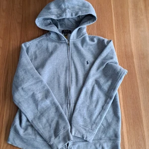 Grå hoodie från Ralph Lauren - Säljer en stilren grå hoodie från Ralph Lauren med dragkedja och broderad logga på bröstet. Perfekt för en avslappnad look. Den har en klassisk passform och är tillverkad i mjukt material för extra komfort. 