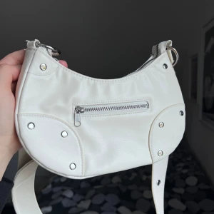 Vit axelväska med dragkedja - Snygg vit axelväska i syntetmaterial med dragkedja och nitar som detaljer. Väskan har en justerbar rem och en praktisk ytterficka med dragkedja. Perfekt för att bära dina nödvändigheter med stil. Väskan har ett stort fack inuti samt ett litet på utsidan. Från H&M. Storlek: 24x12cm