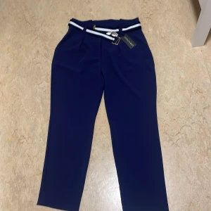 Mörkblå kostymbyxor med bälte - Snygga mörkblå kostymbyxor från LCW modest med ett stilrent bälte i vitt och blått. Byxorna har en rak passform och är perfekta för en elegant look. De har en klassisk design med hällor för bältet och en diskret dragkedja framtill.De är aldrig använda och har kvar prislapp. Köpta i ett annat land för cirka 700kr. Säljer för 450kr. Storlek 42 men passar även som storlek 40.