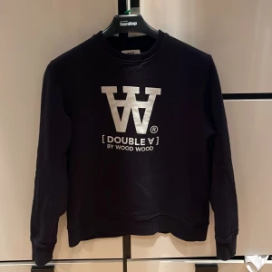 Svart tröja från Wood Wood - Säljer en svart tröja från Wood Wood med tryck 'Double A' på framsidan. Tröjan har långa ärmar och en klassisk rund halsringning. Perfekt för en stilren look. Använd fåtal gånger så är bra skick. Säljer pga den är lite mer baggy style och samtidigt inte. Nypris 1400