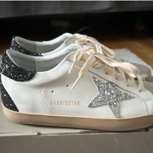 Vita sneakers med glittrig stjärndetalj - Snygga vita sneakers från Golden Goose med en glittrig stjärna på sidan och glittrig häl. Skorna har snörning och en bekväm sula. Perfekta för att ge en touch av glamour till din outfit. Första bilden är från när skorna var nya. Nu är de i använt skick så något smutsiga men inga skador. Nypris är runt 7000kr därav priset men skicka gärna prisförslag om priset inte passar er. Försöker rengöra skorna innan jag postar vid försäljning❤️de är äkta men har tyvärr inget bevis