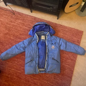 Blå jacka från Moncler - Säljer min snygga Moncler jacka. Har lagat den lite tex är luvan fast sydd eftersom dragkedjan var trasig och det syns knappt. Sen har jag sytt lite på ärmen som ni kan se på sista bilden. Den har små fläckar på framsidan längst ner men syns inte så mycket. En ficka är trasig inuti men kan laga lätt. Det står XXL men tror det är barnstorlek så passar mig som är XS.