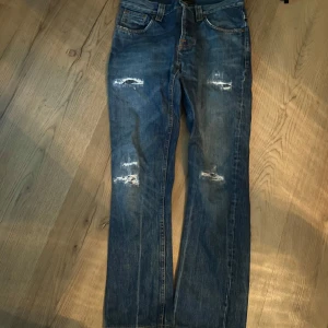 Blå jeans från Nudie Jeans - Säljer ett par blå jeans från Nudie Jeans med slitna detaljer på framsidan. De har en klassisk femficksdesign och är högmidjade med en straight passform. Perfekta för en avslappnad stil.