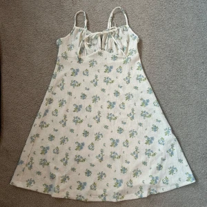 Blommig kort klänning - Charmig blommig klänning med smala justerbara axelband och knytband framtill. Klänningen har ett vackert blommönster i blått och grönt på en vit bakgrund. Perfekt för sommarens varma dagar.