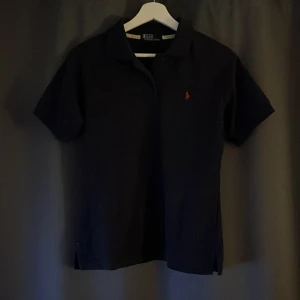Svart pikétröja från Ralph Lauren - Snygg svart pikétröja från Ralph Lauren med klassisk krage och korta ärmar. Den har en röd broderad logotyp på bröstet som ger en stilren touch. Perfekt för en avslappnad men ändå elegant look.