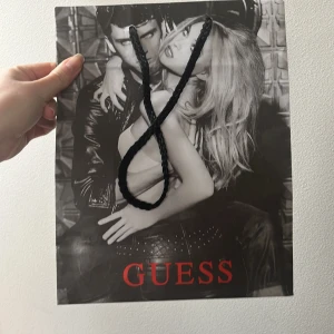 Shoppingväska från Guess - Snygg shoppingväska från Guess med svartvita bilder och röd logotyp. Väskan har svarta flätade handtag och en stilren design. Perfekt för att bära med sig mindre inköp eller som en trendig accessoar.