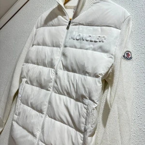 Vit dunjacka från Moncler - Säljer en stilren vit dunjacka från Moncler med dragkedja och broderad logotyp på ärmen. Jackan har en quiltad design framtill och är tillverkad i en kombination av syntet och stickat material. Perfekt för kyligare dagar med sin långa ärmar och bekväma passform. Taggar och lapp samt påse från Moncler medföljer