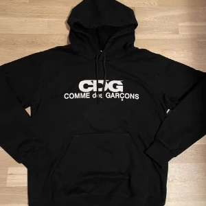 Svart hoodie från Comme des Garçons - Säljer en svart hoodie från Comme des Garçons med vit logga på bröstet. Den har en klassisk design med dragsko i huvan och en stor magficka. Perfekt för en stilren och bekväm look.