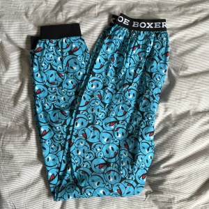 Blå byxor med smiley-mönster från Joe Boxer - Coola blå byxor från Joe Boxer med ett lekfullt smiley-mönster. De har en elastisk midja med märkeslogga och är tillverkade i 100% bomull för extra komfort. Perfekta för en avslappnad stil.