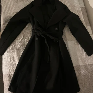 Svart kappa med knytband - Elegant svart kappa med knytband i midjan och klassisk krage. Finns även två jättesnygga knappar på den. Aldrig använd, dvs i helt NYTT SKICK!!!! Storlek M. 