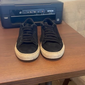 Svarta sneakers från Axel Arigato - Snygga svarta sneakers från Axel Arigato med vit sula och diskret logga på sidan. Skorna har en klassisk design med snörning och är perfekta för en stilren look. Nypris 2500