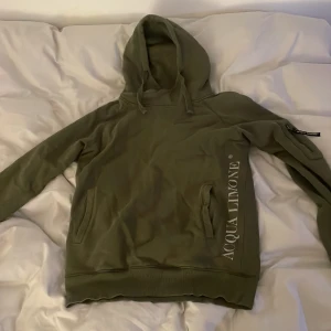Olivgrön hoodie från Acqua Limone - Säljer en olivgrön hoodie från Acqua Limone med deras ikoniska logga på bröstet och ryggen. Tröjan har en bekväm passform med långa ärmar och en praktisk ficka framtill. Perfekt för en avslappnad stil.