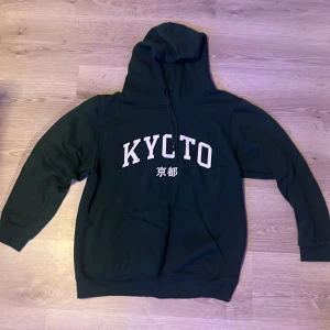 Grön hoodie från boohooMAN - Säljer en svart hoodie från boohooMAN med texten 'KYOTO' i vitt på framsidan. Tröjan har en klassisk design med huva och långa ärmar. Perfekt för en avslappnad stil.