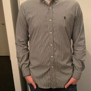 Randig skjorta från Ralph Lauren - Snygg randig skjorta från Ralph Lauren i klassisk passform. Skjortan är svart och vit med långa ärmar och knappar framtill. Perfekt för en stilren look.