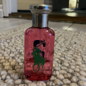 Rosa parfym från Ralph Lauren - Säljer en rosa parfym från Ralph Lauren med en grön polospelare och en stor blå siffra 2 på framsidan. Flaskan har en silverfärgad kork och en stilren design. Perfekt för den som gillar en sportig och fräsch doft. Har använt ca 2 tredjedelar