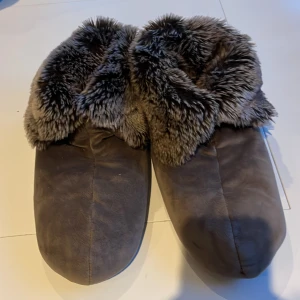 Grå tofflor med fuskpäls från Newport - Mysiga brun grå tofflor från Newport med mjuk fuskpäls runt kanten. Perfekta för kalla dagar hemma. De har en stabil sula och är gjorda i ett syntetmaterial som ger extra komfort.