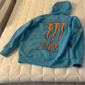 Blå hoodie från Monster Energy - Säljer en blå hoodie från Monster Energy med deras ikoniska logga i orange på både fram- och baksidan. Tröjan har en stor ficka framtill och långa ärmar med tryck på. Perfekt för en avslappnad stil.
