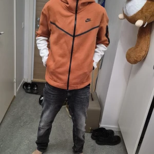 Orange hoodie från Nike - Säljer en snygg orange hoodie från Nike med svarta detaljer och dragkedja. Jackan har en huva och en cool ficka med dragkedja på ärmen. Perfekt för en sportig look!