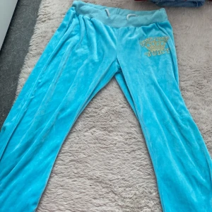 Turkosa mjukisbyxor från Juicy Couture - Snygga turkosa mjukisbyxor från Juicy Couture med guldbroderad text och krona på sidan. Byxorna har en bekväm resår i midjan och är tillverkade i ett mjukt material som ger en lyxig känsla. Perfekta för en avslappnad stil.