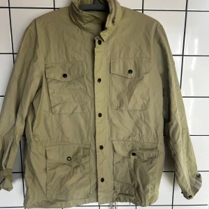 Beige jacka från Fjällräven - Säljer en beige jacka från Fjällräven med knappar och flera praktiska fickor. Jackan har en klassisk design med långärmade ärmar och en broderad logga på ärmen. Perfekt för utomhusaktiviteter och vardagsbruk. Luva finns infällt i kragen. 