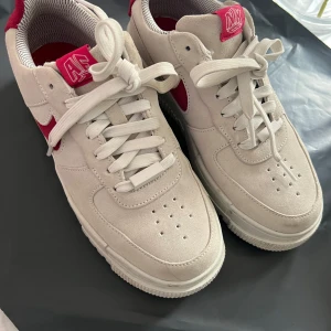 Nike Air Force i beige och röd - Snygga Nike Air Force sneakers i beige mocka med röda detaljer. Skorna har klassisk snörning och en robust sula med AIR-dämpning för extra komfort. Perfekta för en stilren look. Storlek 38.5