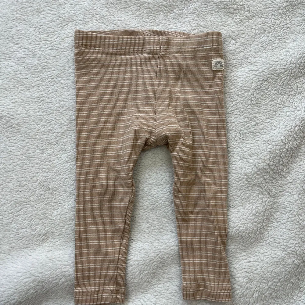 Säljer ett par söta beige och vita randiga leggings för baby från Lindex. De är mjuka och bekväma, perfekta för de minsta. Leggingsen har en liten etikett med en regnbåge på sidan.. Farkut & Housut.
