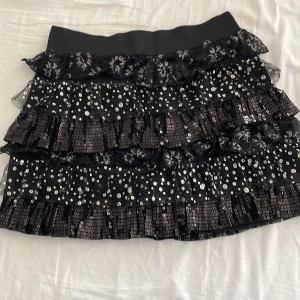 Svart glittrig kjol med volanger - Snygg svart kortkjol med glittriga paljetter och flera lager av volanger. Perfekt för att sticka ut med en festlig look. Kjolen har en elastisk midja för bekväm passform. Står ej storlek men skulle säga S/M