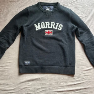 Svart tröja från Morris - Snygg svart tröja från Morris med vit text och en broderad brittisk flagga på bröstet. Tröjan har långa ärmar och ribbade muddar vid ärmslut och nederkant. Perfekt för en stilren look. Storlek L men passar M